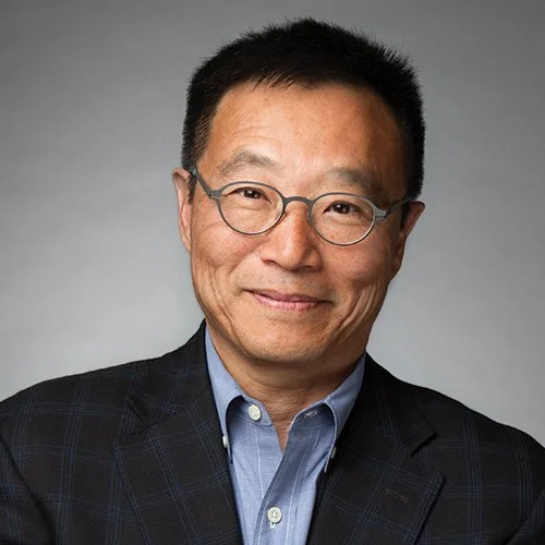 A man with short black hair wearing glasses, a dark blazer, and a light blue collared shirt smiling at the camera against a gray background.
