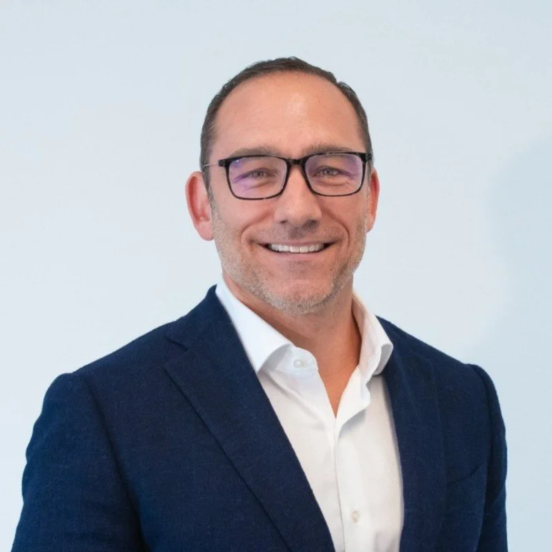 A middle-aged man with glasses, wearing a white shirt and dark blazer, smiling in front of a plain light-colored background.