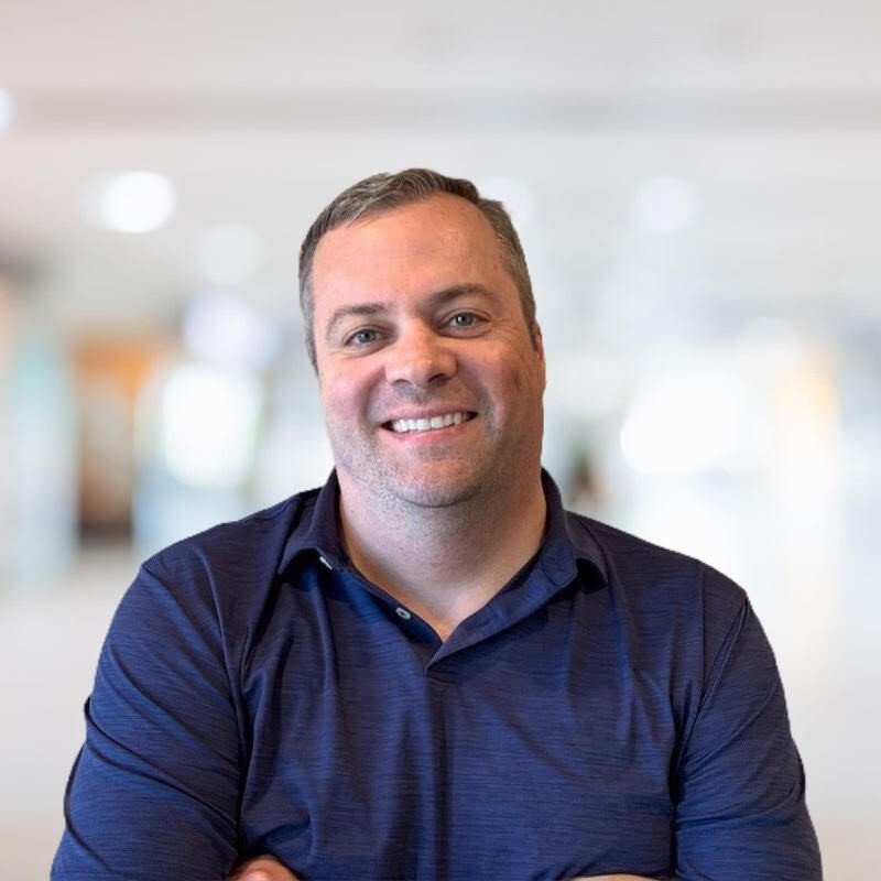 A smiling man with short brown hair wearing a dark blue collared shirt, standing with arms crossed in an indoor, well-lit environment.
