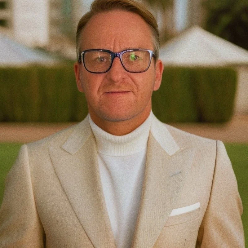 A man in a beige suit and white turtleneck sweater standing outdoors, wearing glasses, with a neutral expression and a background of bushes and tents.