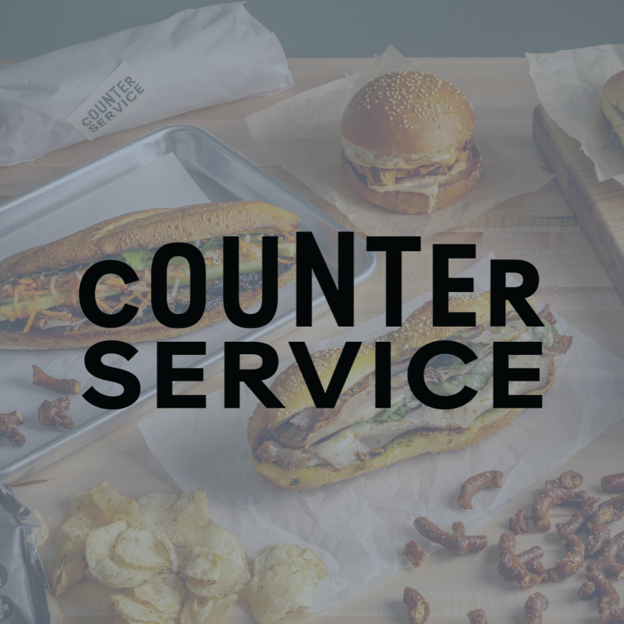 Fast food meal including a sandwich on sandwich paper, a burger with sesame bun, fried items like onion rings and crispy bacon bits, all on a wooden table