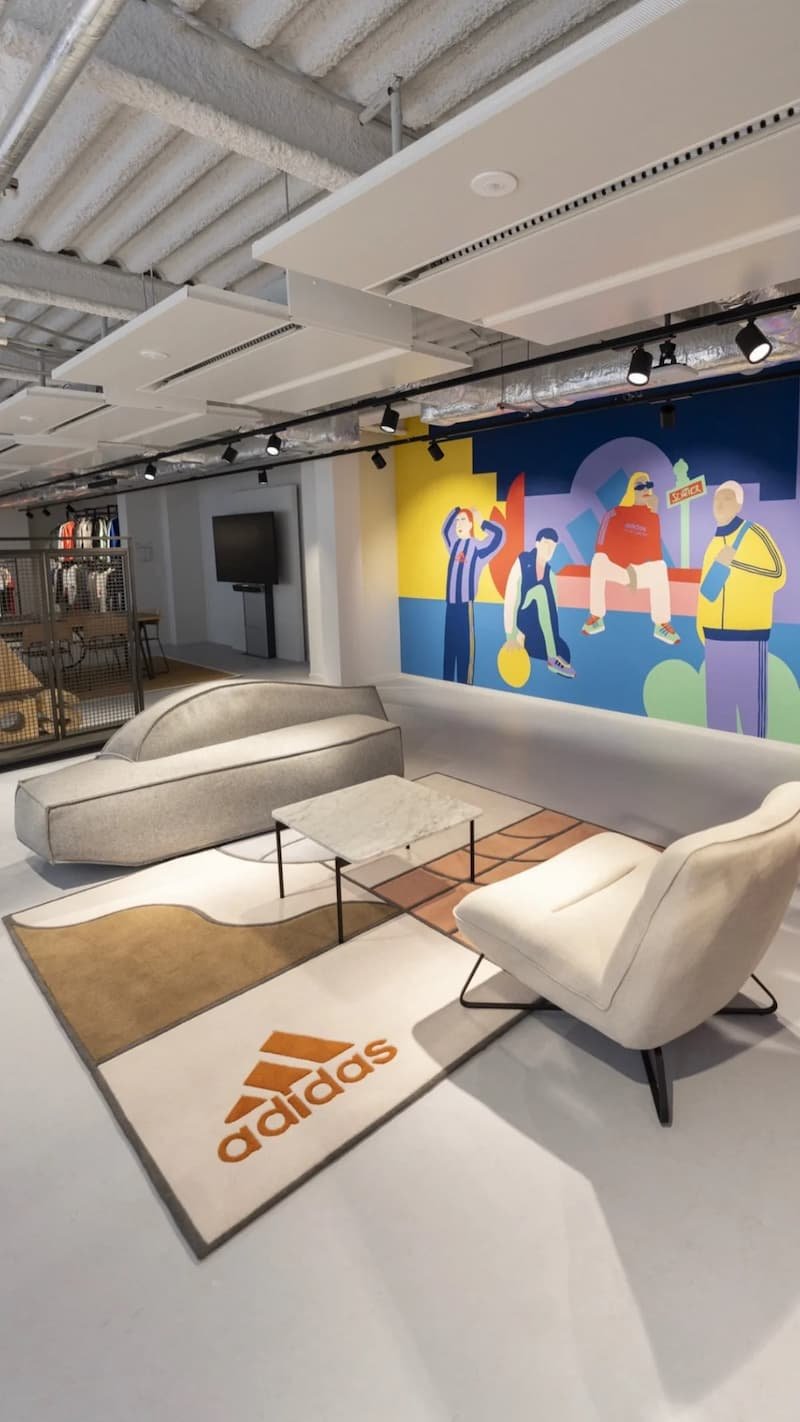 A lounge featuring an Adidas rug, two armchairs, a coffee table, a wall with a colorful mural depicting several people in an urban style, a flat-screen TV, and ceilings under construction with lighting fixtures installed.