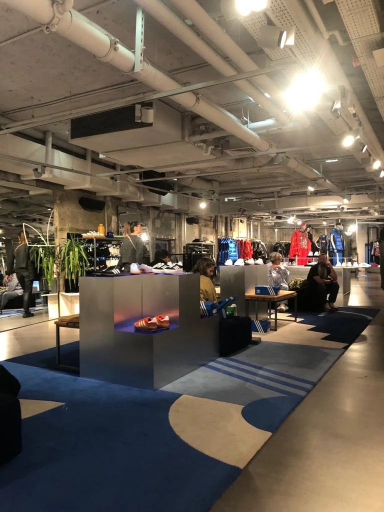 Intérieur d'une boutique de vêtements avec plusieurs clients assis et en train de magasiner, portant des vêtements colorés et des manteaux exposés à l'arrière.