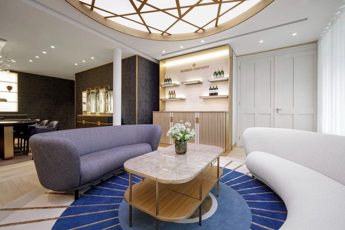 An elegant living room featuring a grey sofa, a white armchair, a marble coffee table, and shelves filled with bottles, all within a bright and contemporary space.