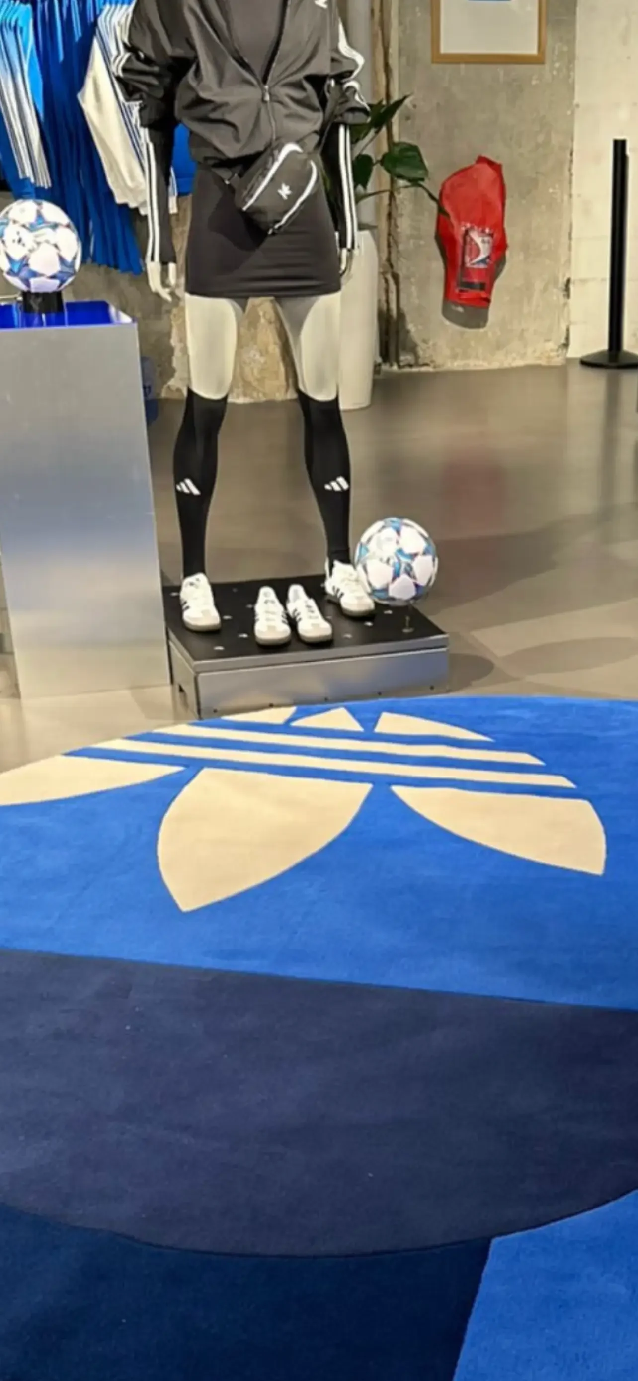 Mannequin habillé en vêtements de sport avec deux ballons de football, chaussures blanches et sac à bandoulière, dans une boutique, avec un tapis bleu et blanc au sol.