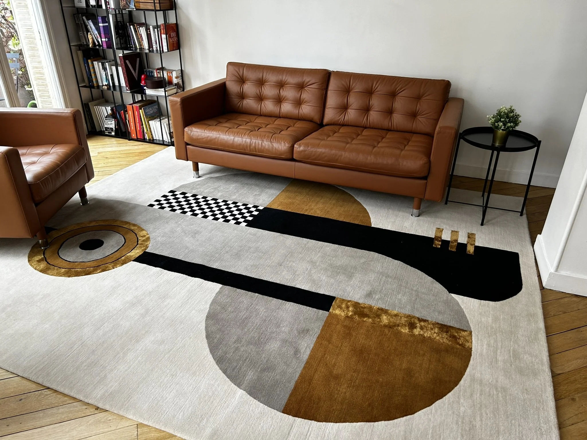 Salon lumineux avec canapé en cuir marron, fauteuils en cuir assortis, tapis coloré avec formes géométriques, étagère noire avec livres, petite table ronde avec une plante verte, parquet en bois.