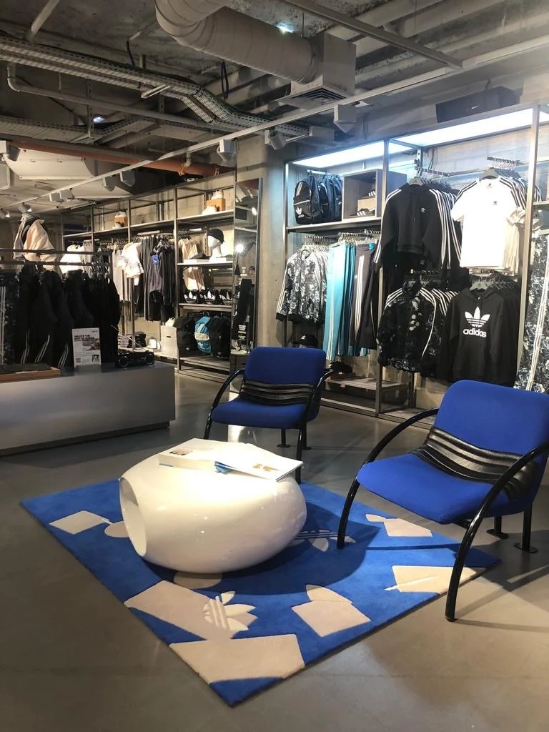 Coin salon dans un magasin de vêtements avec deux chaises bleues, une table blanche, un tapis bleu et des vêtements de sport exposés derrière.
