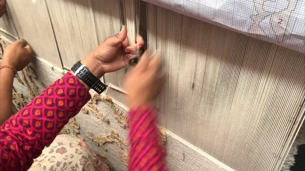 A rug depicting two people assembling warp threads on a wooden loom, preparing for hand-knotting.