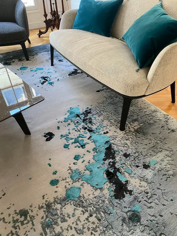 Un tapis moderne avec des taches de peinture bleu, noir et gris dans un salon avec canapé beige et coussins bleus.