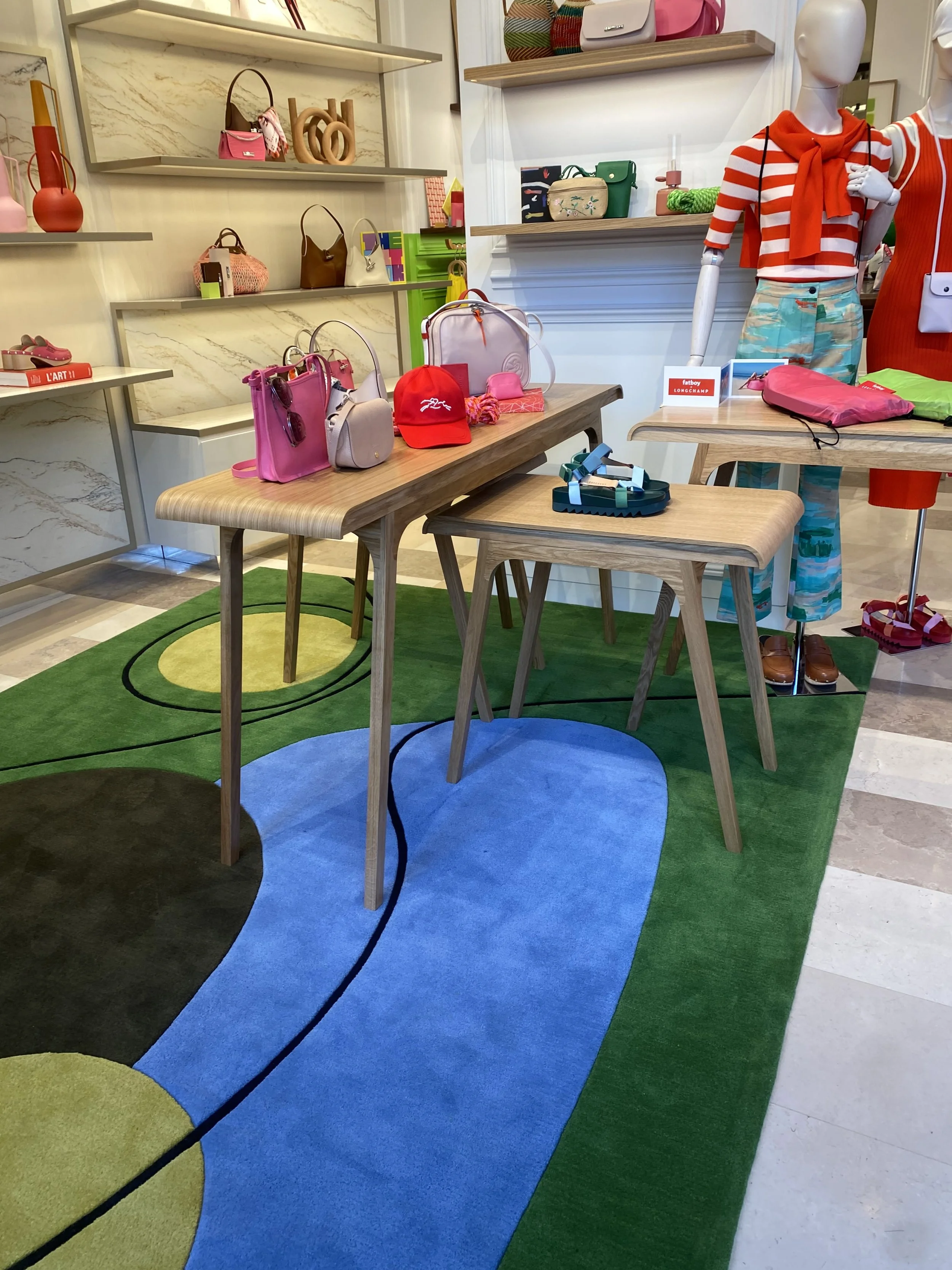 Vitrine de boutique avec des sacs à main colorés, un mannequin habillé en vêtements à rayures rouges et blanches, portant une robes à rayures, une table en bois avec des accessoires, et un tapis avec design coloré.