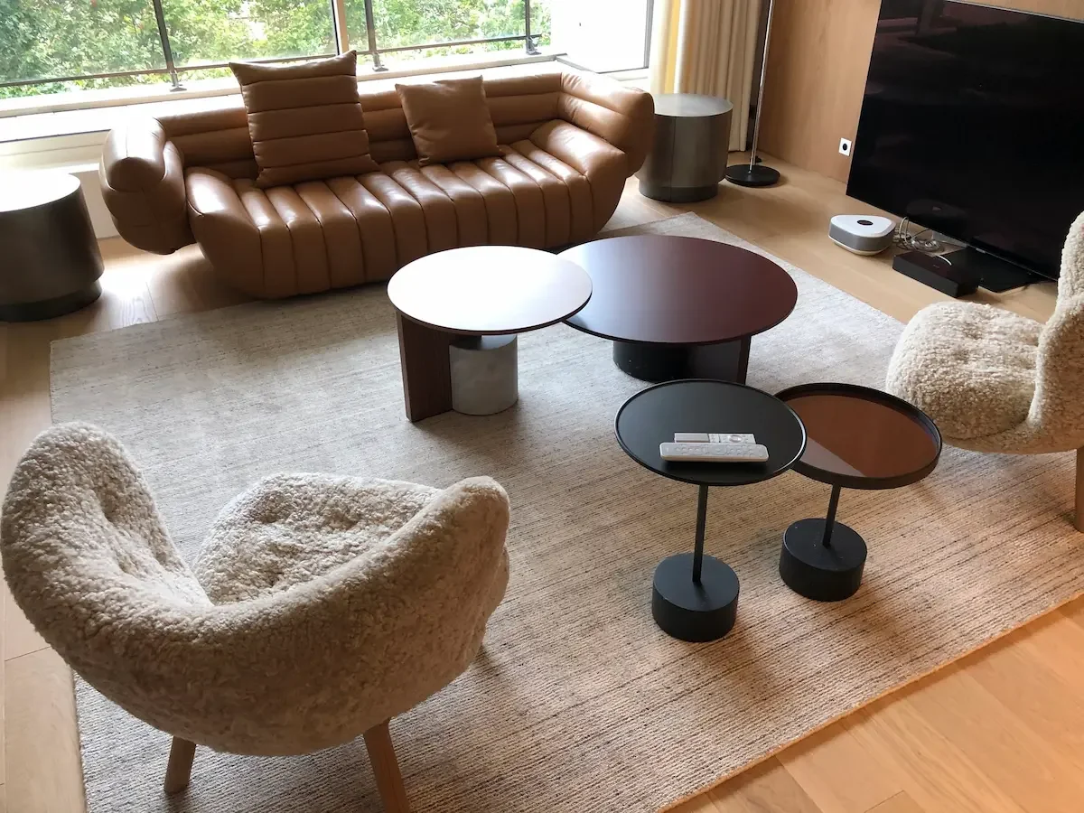 A living room featuring a light brown leather sofa, two beige fabric armchairs, dark wood coffee tables, a radiator, a large-screen TV, a beige rug on a wooden floor, and a large window overlooking trees.