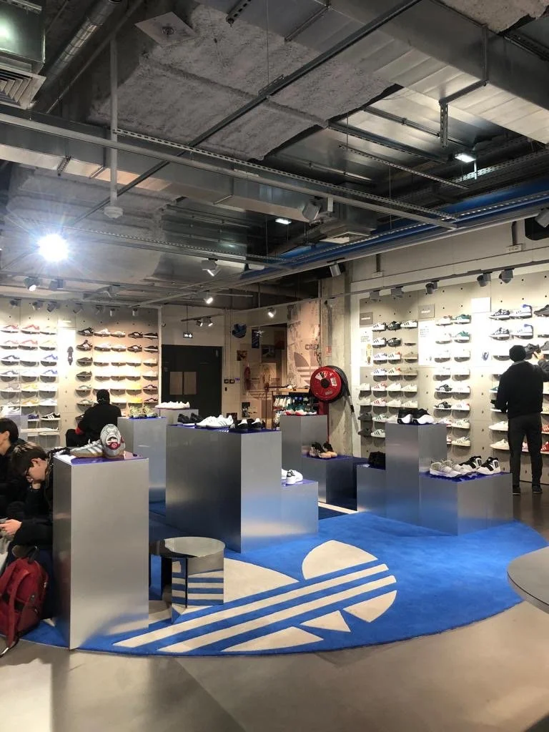 Magasin de chaussures avec un mur de chaussures à l'étage, plusieurs personnes essayant ou regardant les chaussures, sol en béton et plafond industriel avec conduits visibles, tapis bleu avec logo Adidas.