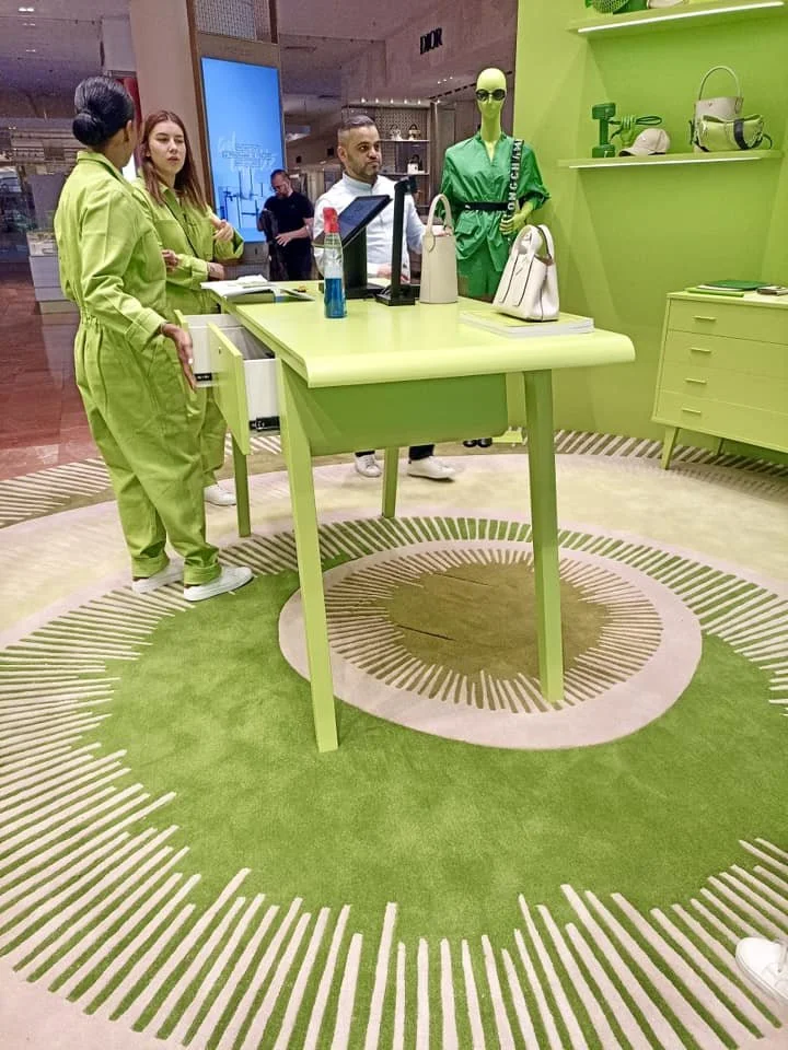 Boutique avec mannequin portant un sac à main et un pantalon vert, personnes discutant au comptoir, décor vert avec meubles et étagères.