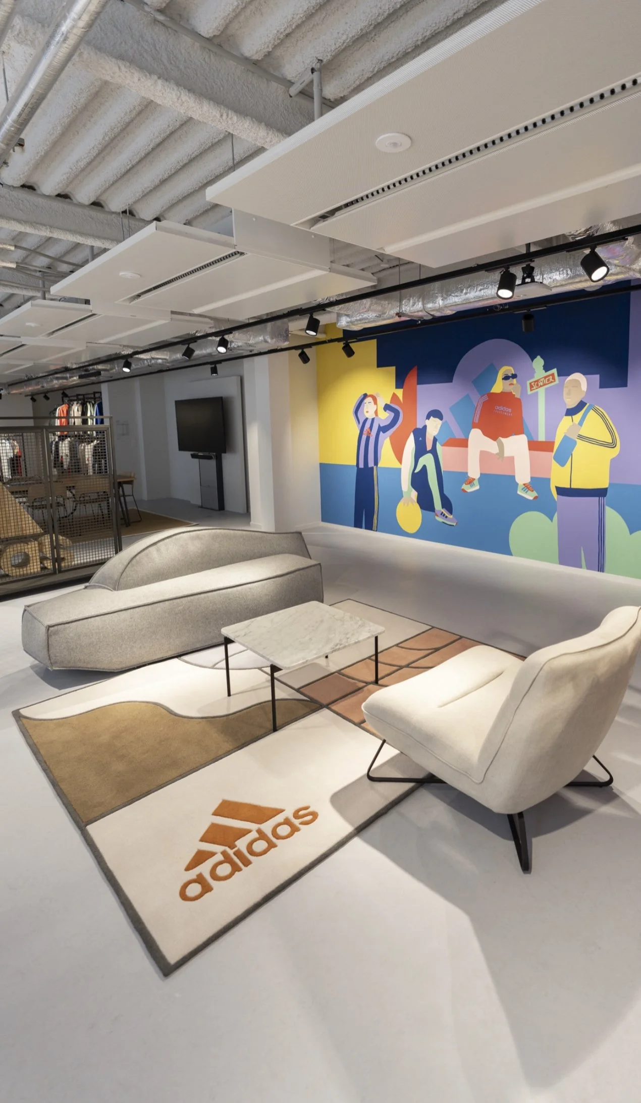 Espace salon avec canapé gris, fauteuil blanc, tapis avec logo adidas, mur coloré avec illustration de jeunes, télévision