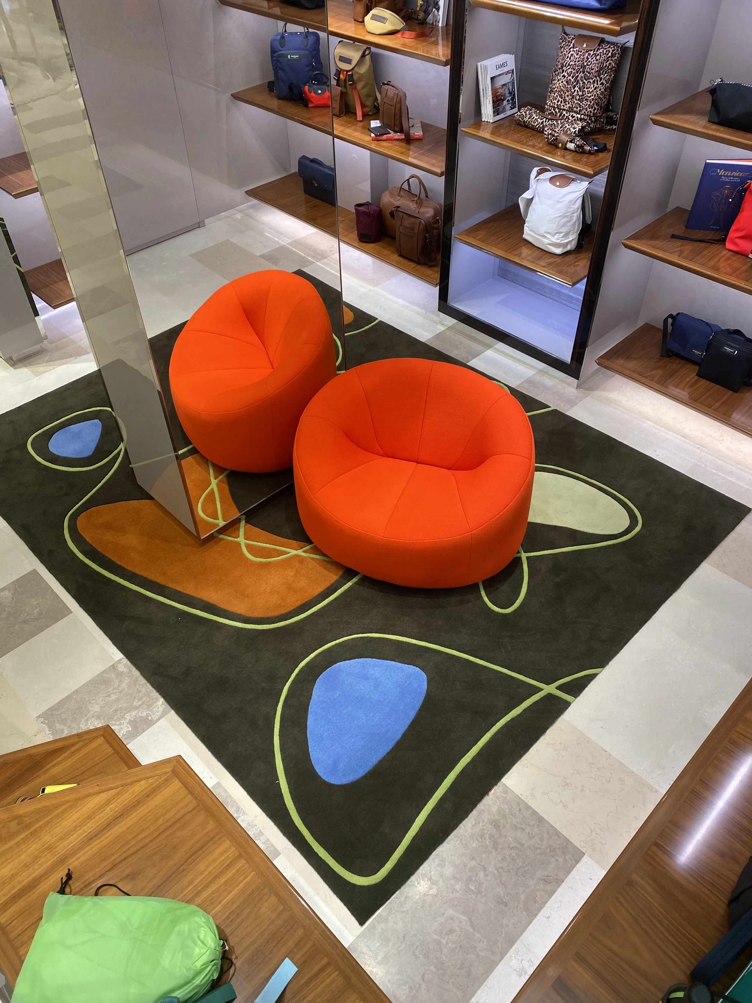 Deux fauteuils orange arrondis placés sur un tapis noir avec des motifs colorés, entourés d'étagères en bois avec des sacs, des livres et des accessoires.