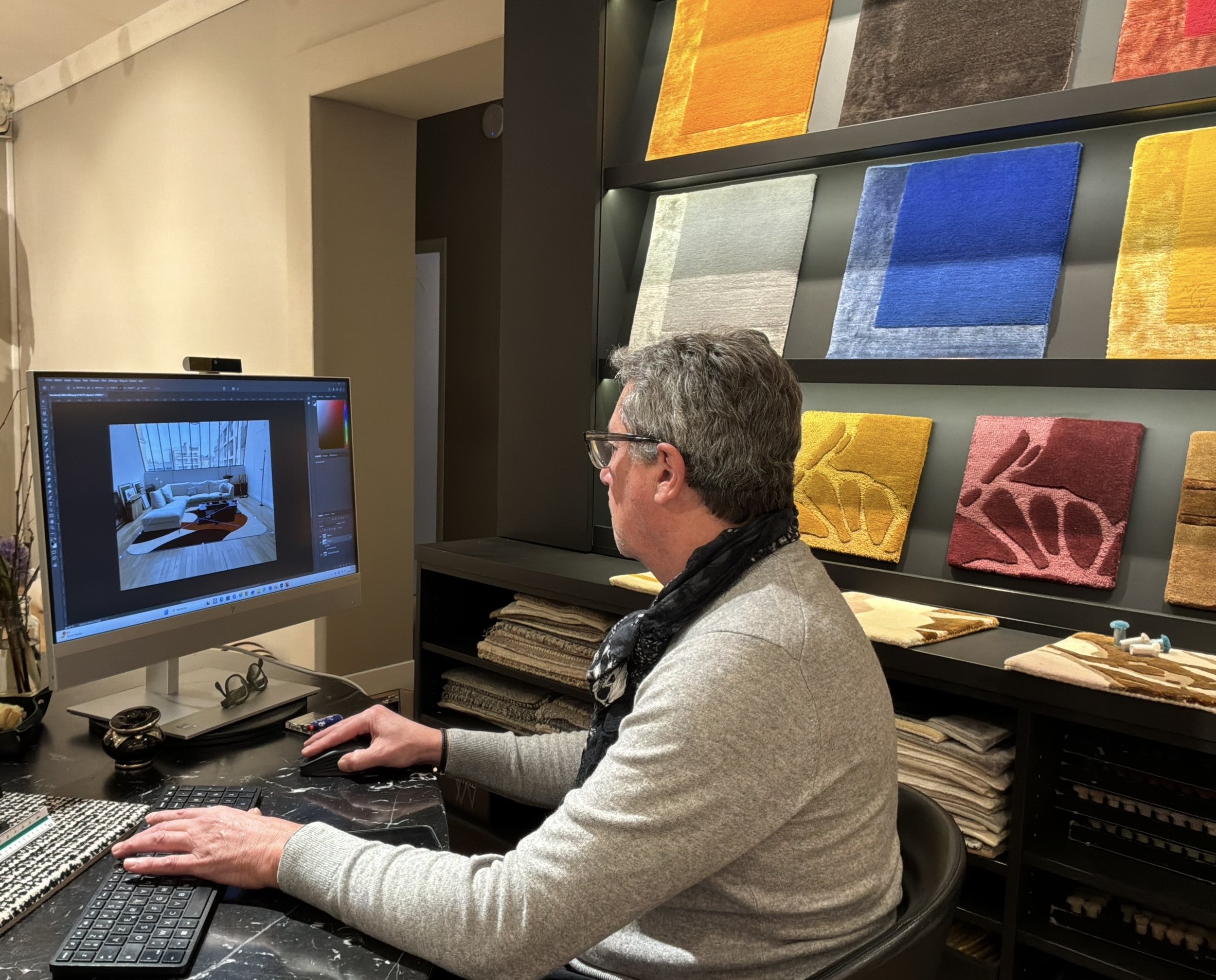 Un homme assis à un bureau travaillant sur un ordinateur, avec un mur de tapis colorés en arrière-plan dans un environnement intérieur.