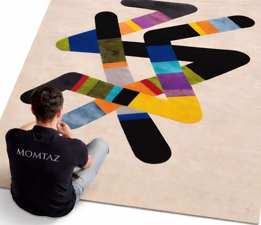 Une personne portant un t-shirt noir avec le mot 'MOMTAZ' écrit dessus, assise sur un sol beige, regarde un tapis coloré, abstrait, avec des formes géométriques vives.