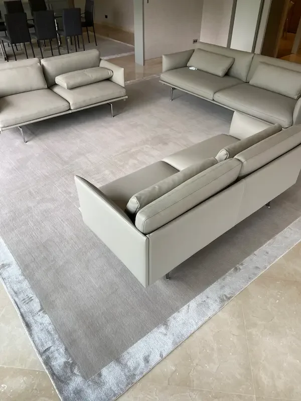 Salon avec trois canapés en cuir gris clair disposés autour d'un tapis gris, dans une pièce moderne.
