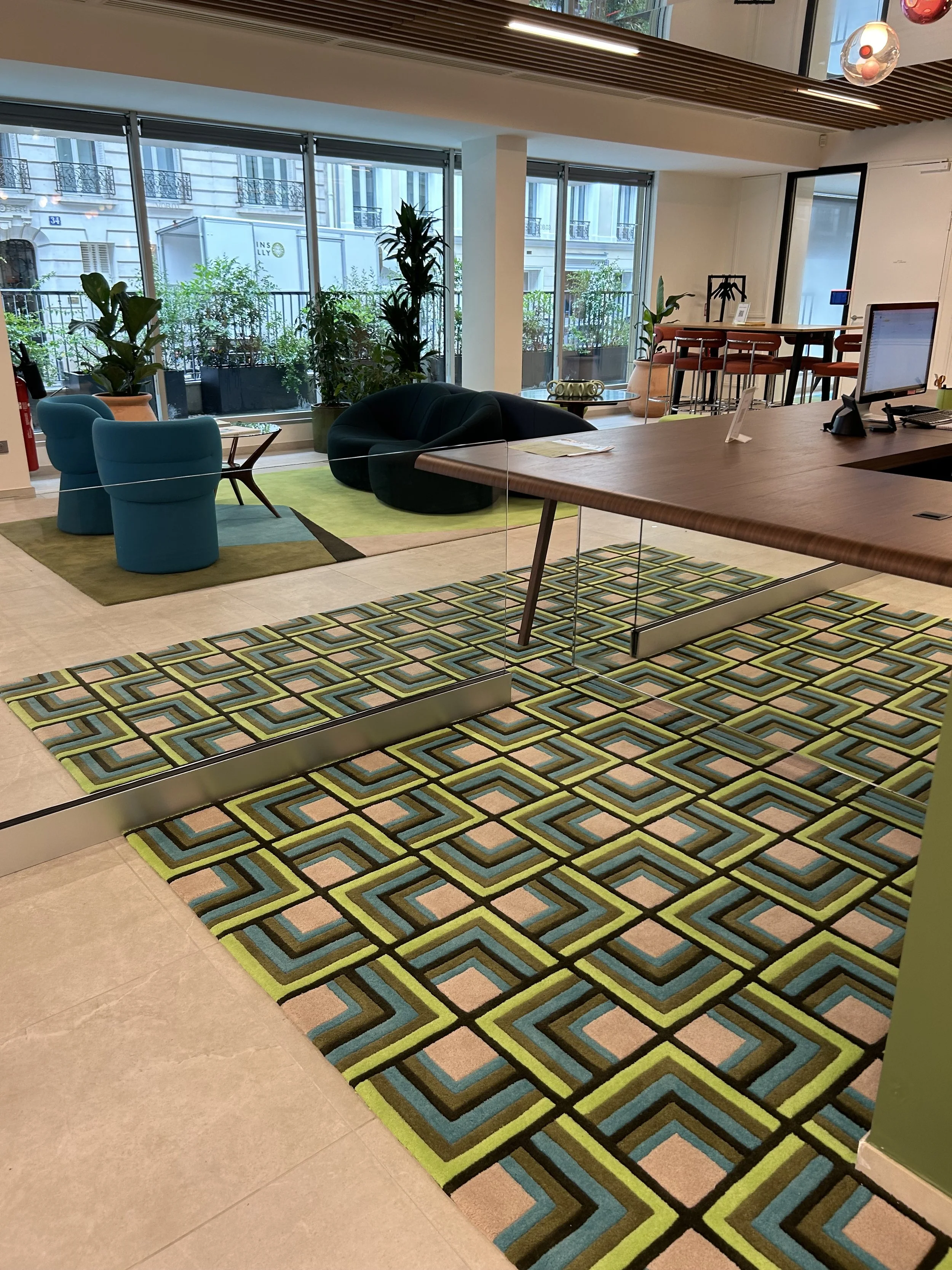 Intérieur moderne avec un tapis géométrique, des fauteuils bleus, un canapé noir, des plantes en pots, et un bureau avec un ordinateur, vue sur une terrasse extérieure avec des fenêtres grandes et lumineuses.