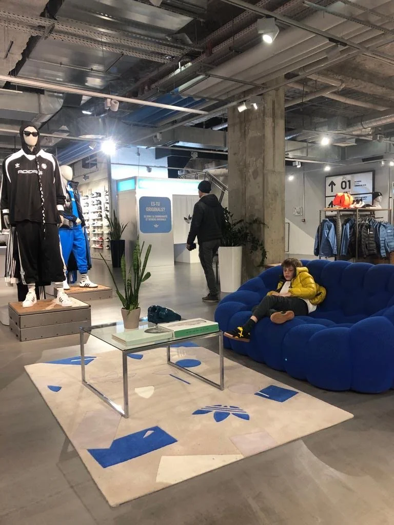 Une boutique de vêtements avec un mannequin portant un survêtement noir et blanc, une étagère avec des vêtements bleus, un canapé bleu où un enfant est assis avec un livre, une table basse avec des livres, et des plantes décoratives.