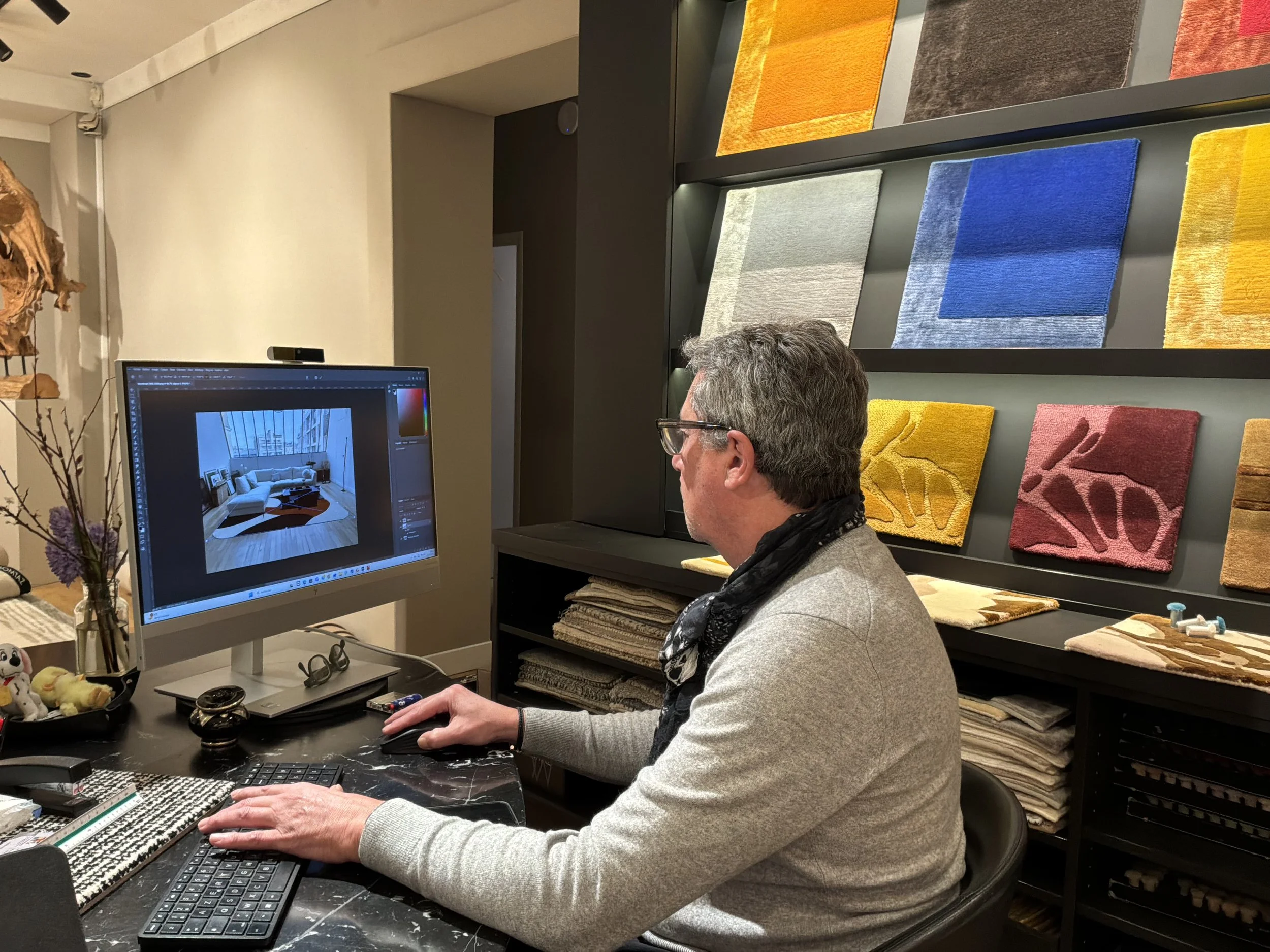 Homme travaillant sur un ordinateur dans un magasin de décoration avec des échantillons de tapis colorés au mur.