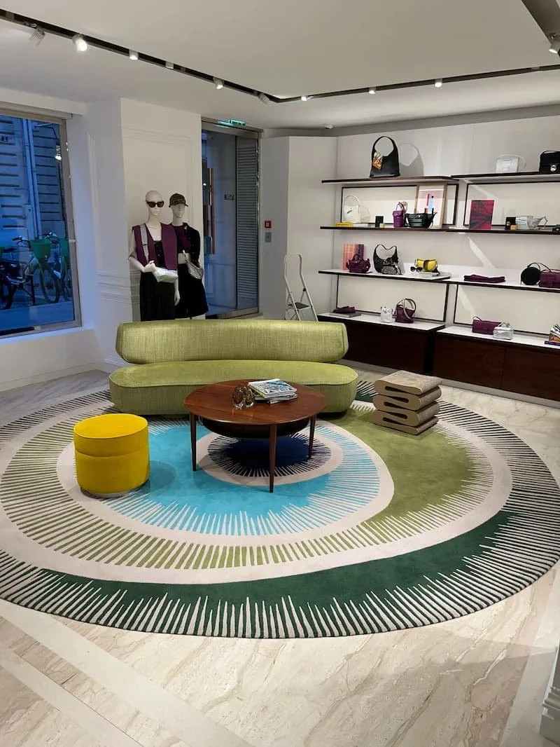 The interior of a boutique featuring dressed mannequins, a green sofa, a round wooden table, a colorful round rug, shelves displaying bags and accessories, and a large window on the left side.