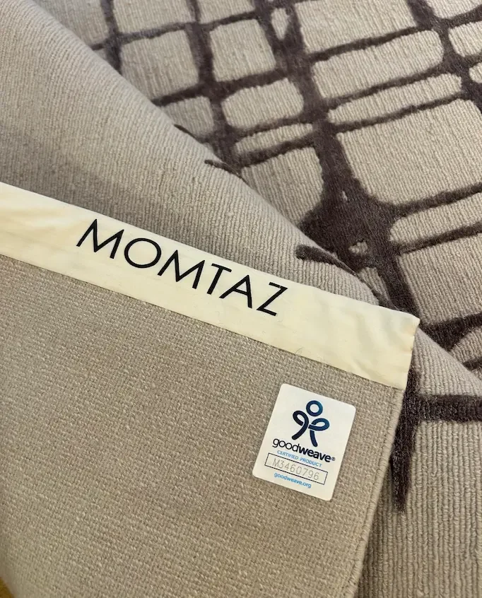 Un tapis beige sur un sol, avec un recyclé de tissu gris clair en laine ou lin. Il y a une étiquette blanche avec le texte "MOMTAZ" et une petite étiquette GoodWeave."
