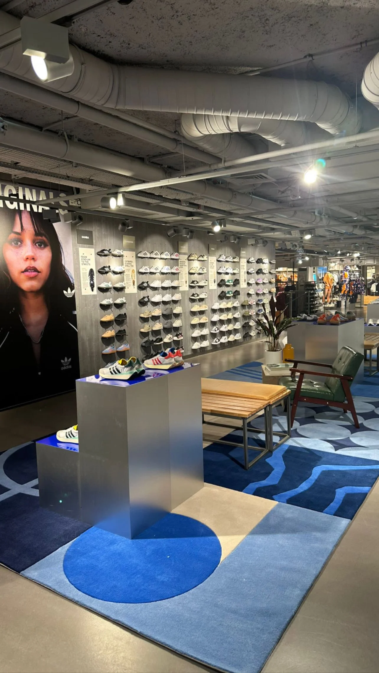 Vitrine de chaussures de sport Adidas dans un magasin de vêtements, avec une grande affiche d'une femme sur le mur et un coin salon avec tapis coloré.