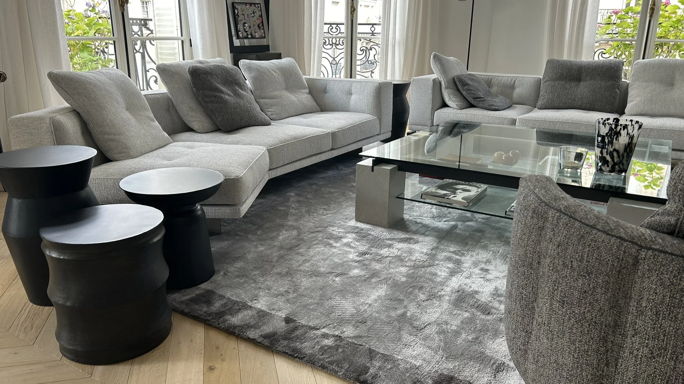Salon moderne avec deux grands canapés gris, une table en verre, trois petites tables noires, un tapis gris et des coussins gris foncés, avec fenêtres donnant sur un balcon avec des plantes vertes