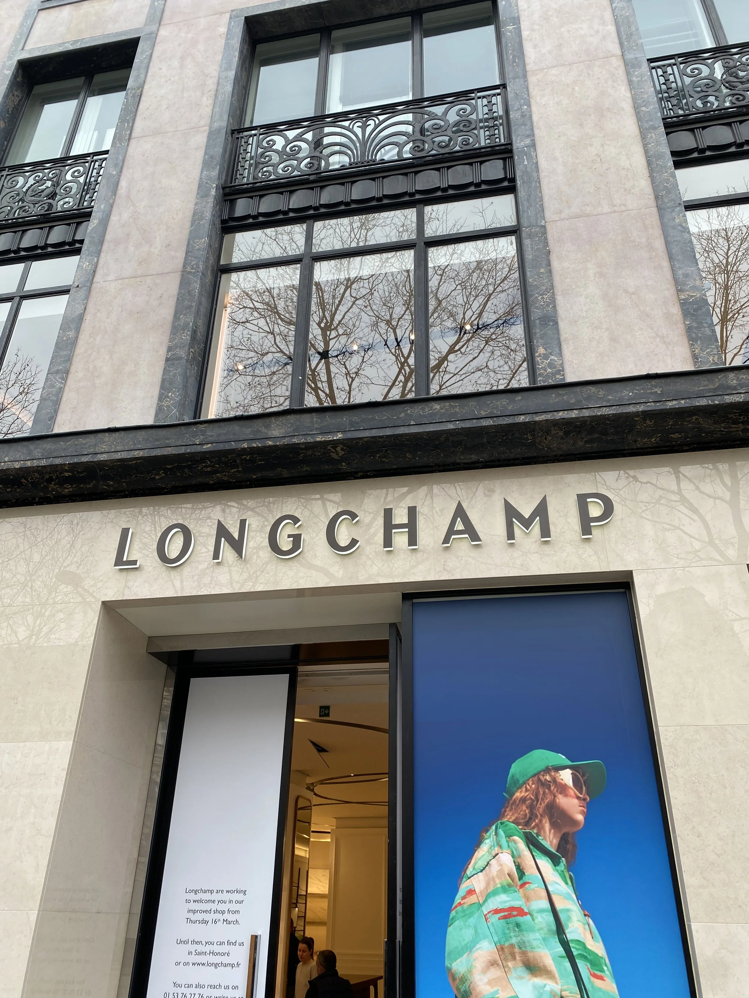 Façade d'un magasin Longchamp avec une grande affiche présentant une femme portant un sac à dos et un chapeau vert, devant une entrée moderne.