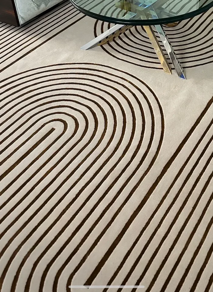Tapis beige avec motifs de lignes courbes brunes, sous une table en verre avec un pied en métal doré et blanc.