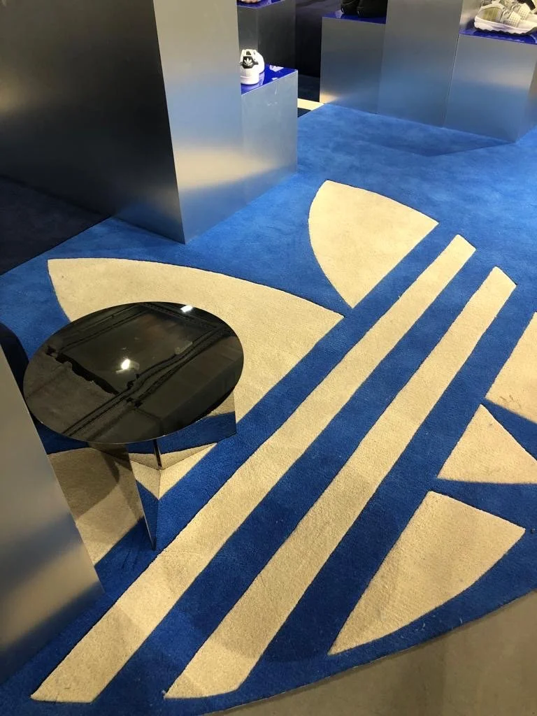 Tapis coloré avec motifs géométriques en bleu et beige, une table noire ronde, présentoirs avec des chaussures dans un espace de magasin.