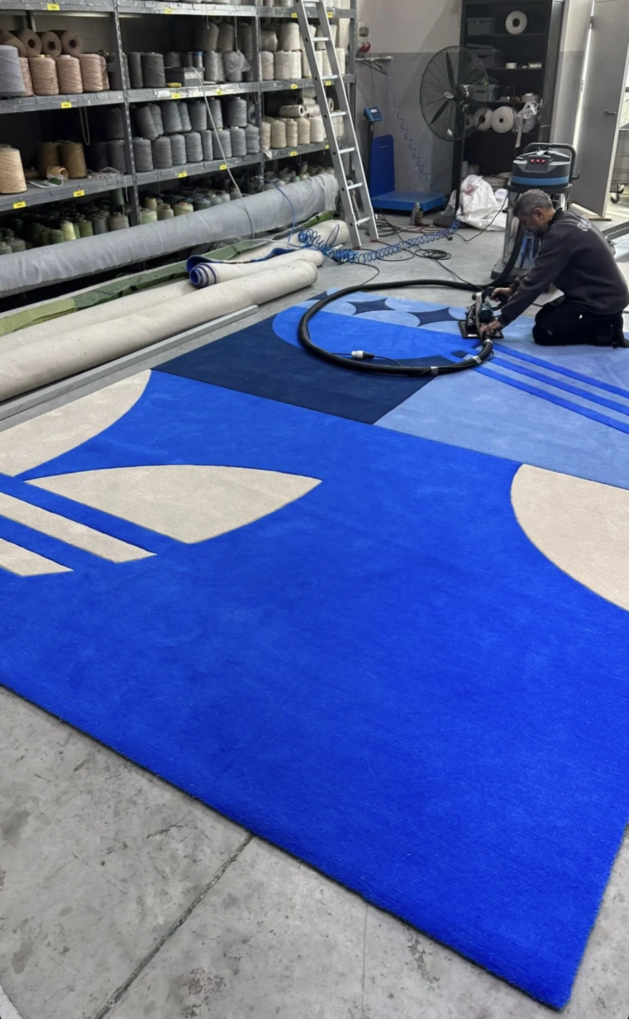 Un homme nettoie ou repasse un grand tapis coloré avec des motifs en bleu, blanc et noir dans un atelier.