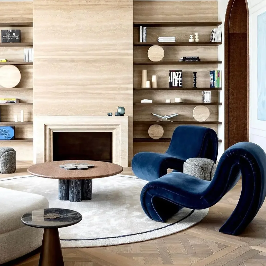 Intérieur d'un salon avec cheminée, étagères en bois avec des livres et décorations, fauteuils en velours bleu, tapis clair, table basse en bois, grande fenêtre laissant entrer la lumière.