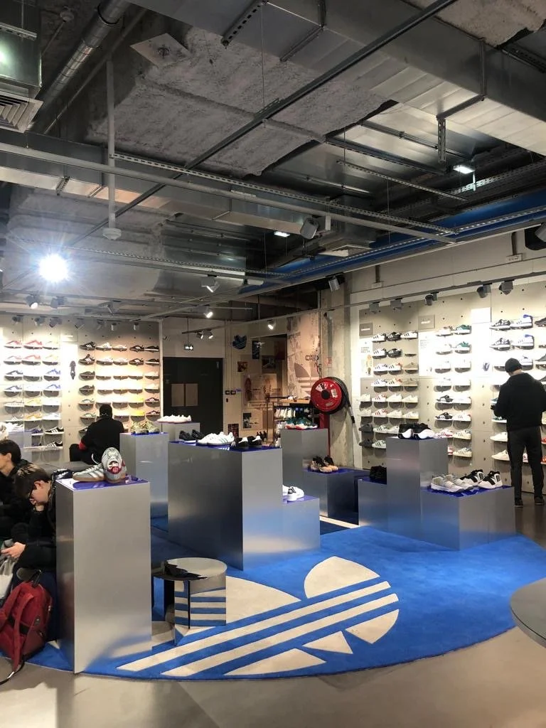 Boutique de chaussures avec un mur de chaussures sur les étagères, plusieurs clients examinant les chaussures, un tapis en forme de logo Adidas, et un plafond industriel avec des éléments exposés.