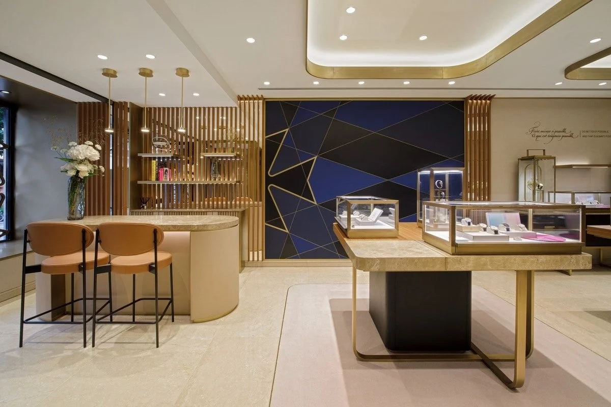 The interior of an elegant jewelry store featuring a marble counter, glass displays containing jewelry, walls decorated in blue and gold, and modern furniture in beige and gold.