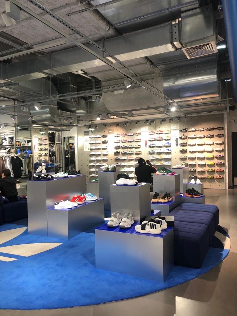 Mur d'étagères avec une collection de chaussures de sport variées dans une boutique. Espace d'exposition avec plusieurs paires de sneakers exposées sur des socles en métal. Quelques clients explorant la sélection, ambiance moderne et organisée.
