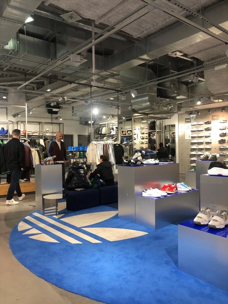 Intérieur d'un magasin de vêtements et chaussures avec plusieurs clients et employés, étagères de vêtements, chaussures sur des présentoirs, un canapé noir avec des personnes assises, tapis rond bleu avec un logo blanc.