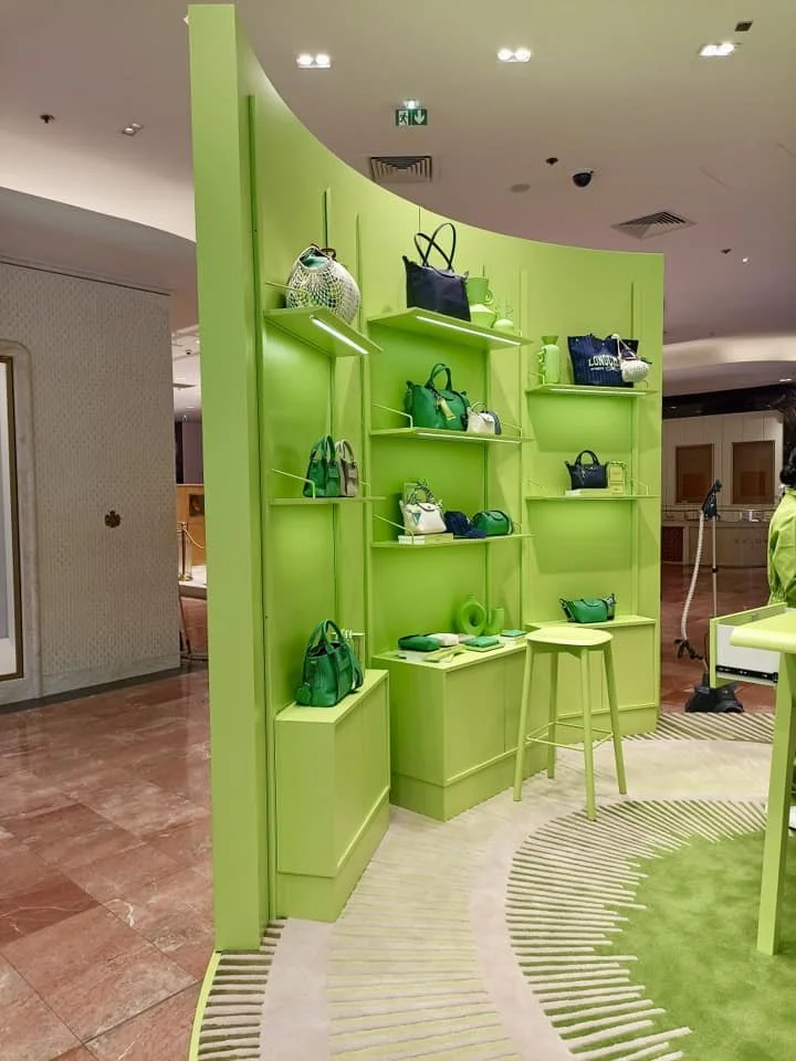 Une display de sacs à main verts dans une boutique, avec un fond de mur vert et un sol en carrelage roseâtre. La mise en scène est minimaliste avec des étagères intégrées.