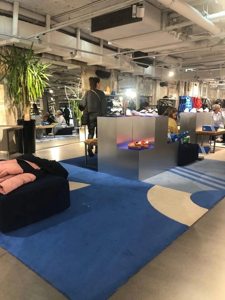 Intérieur d'un magasin de vêtements avec des clients, un mannequin, des vêtements sur des étagères, un canapé avec des vêtements pliés et un tapis.