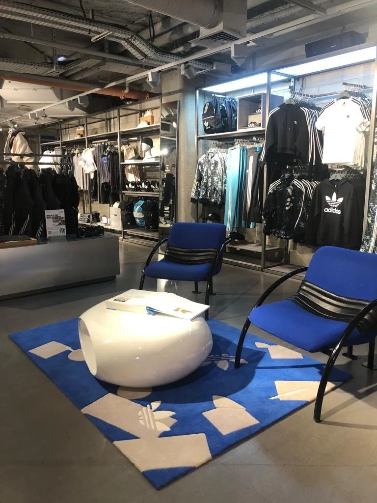Espace salon dans un magasin de vêtements, avec deux fauteuils bleus, une table blanche, un tapis bleu avec motifs, et des vêtements en arrière-plan.
