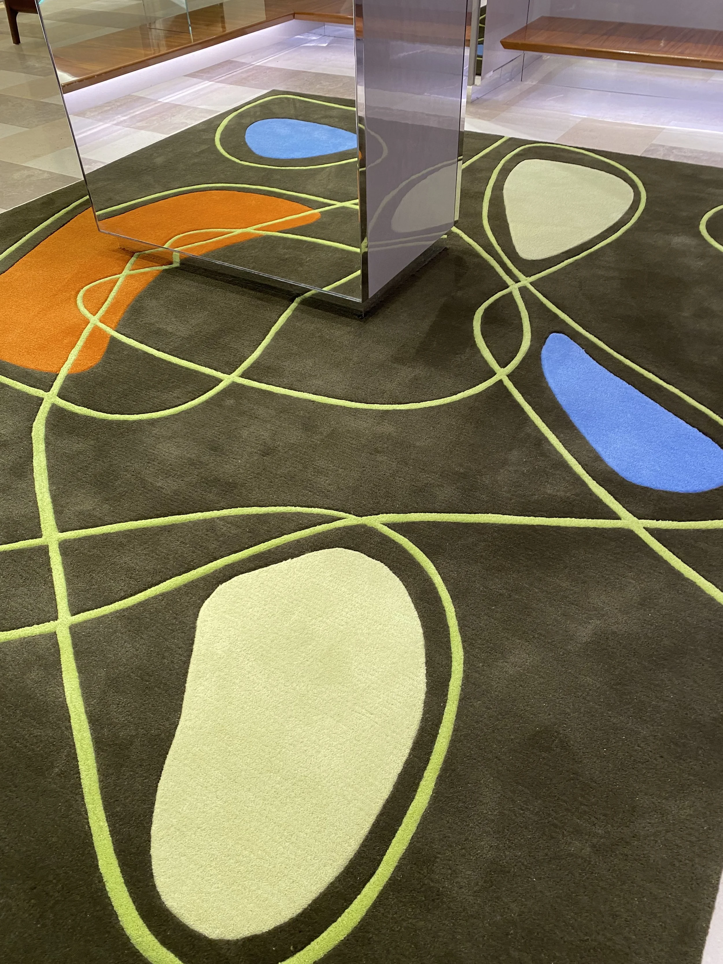 Tapis décoratif dans une zone intérieure, avec des lignes courbes et des formes colorées en bleu, orange, et beige, entourées de lignes vertes.