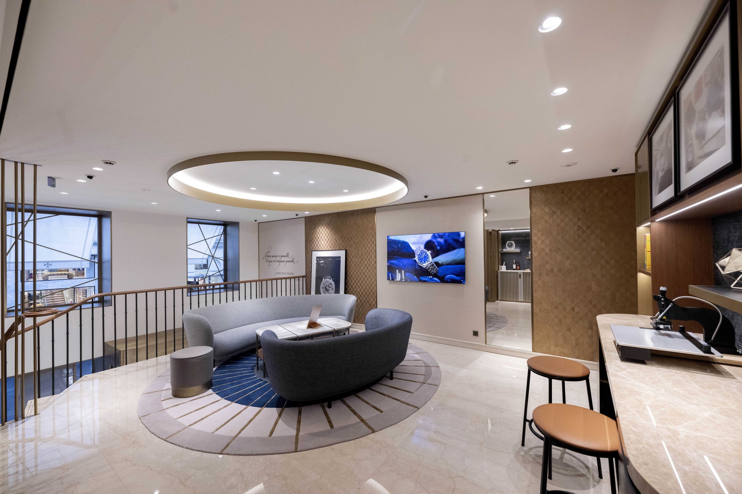 A modern interior featuring a lounge area with a grey sofa, an armchair, a coffee table, a flat-screen TV, a bar with two stools, and a partially visible kitchen in the background.