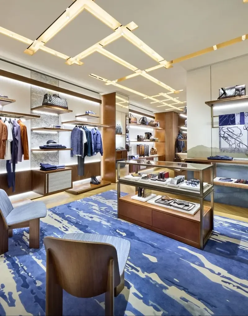 The interior of a clothing and accessories boutique featuring shelves, elegant clothing, handbags, a glass checkout counter, and a blue rug with abstract patterns.