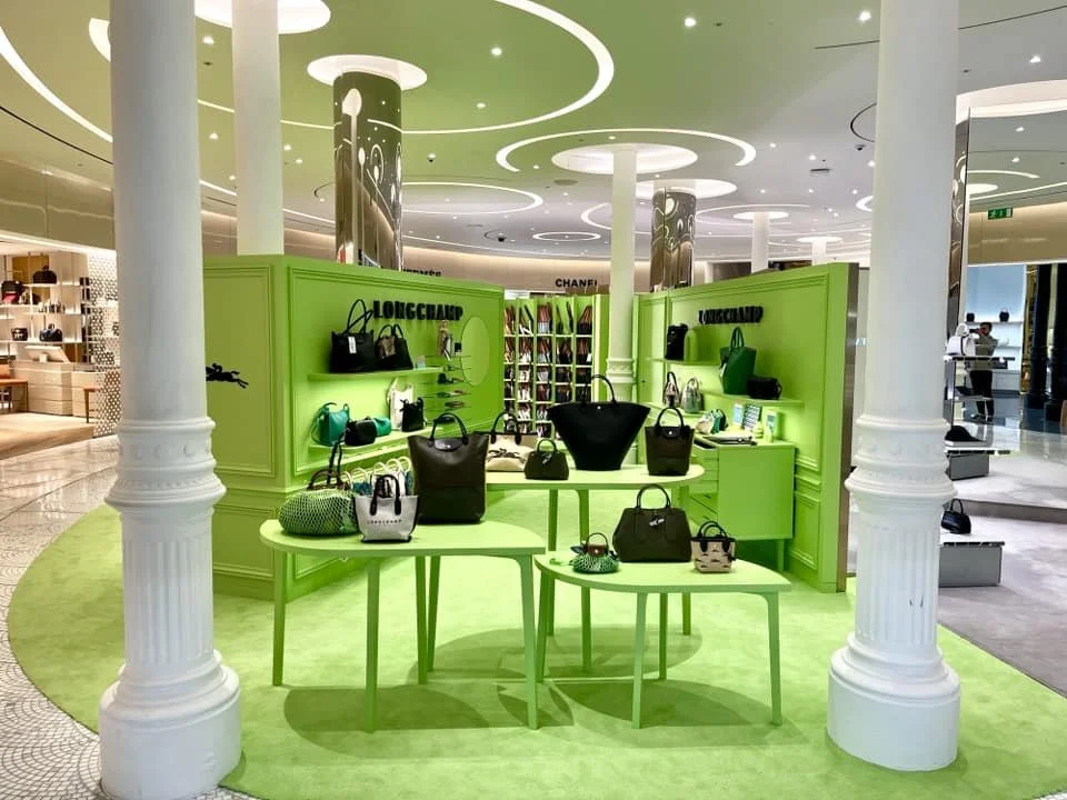 Vitrine de sacs à main de la marque Longchamp dans un centre commercial, avec un décor vert vif et un plafond moderne.