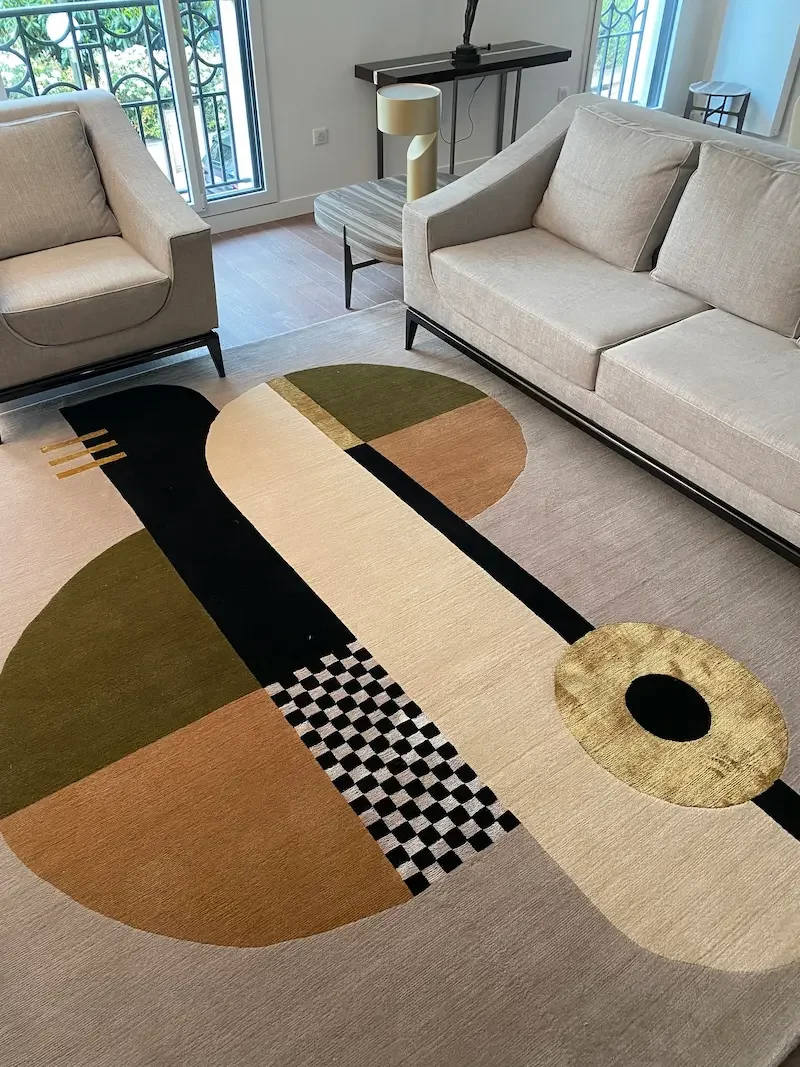 Un salon moderne avec un tapis géométrique aux couleurs de beige, vert, noir, et marron, entouré de deux canapés clairs et une lampe de table, vue d'une fenêtre avec un balcon en arrière-plan.