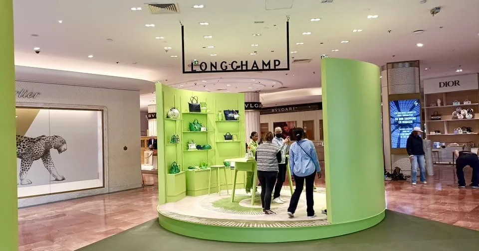 Un stand de marque Longchamp dans un centre commercial, avec des étagères vertes affichant des sacs à main, et des personnes en train de discuter autour d'une table.