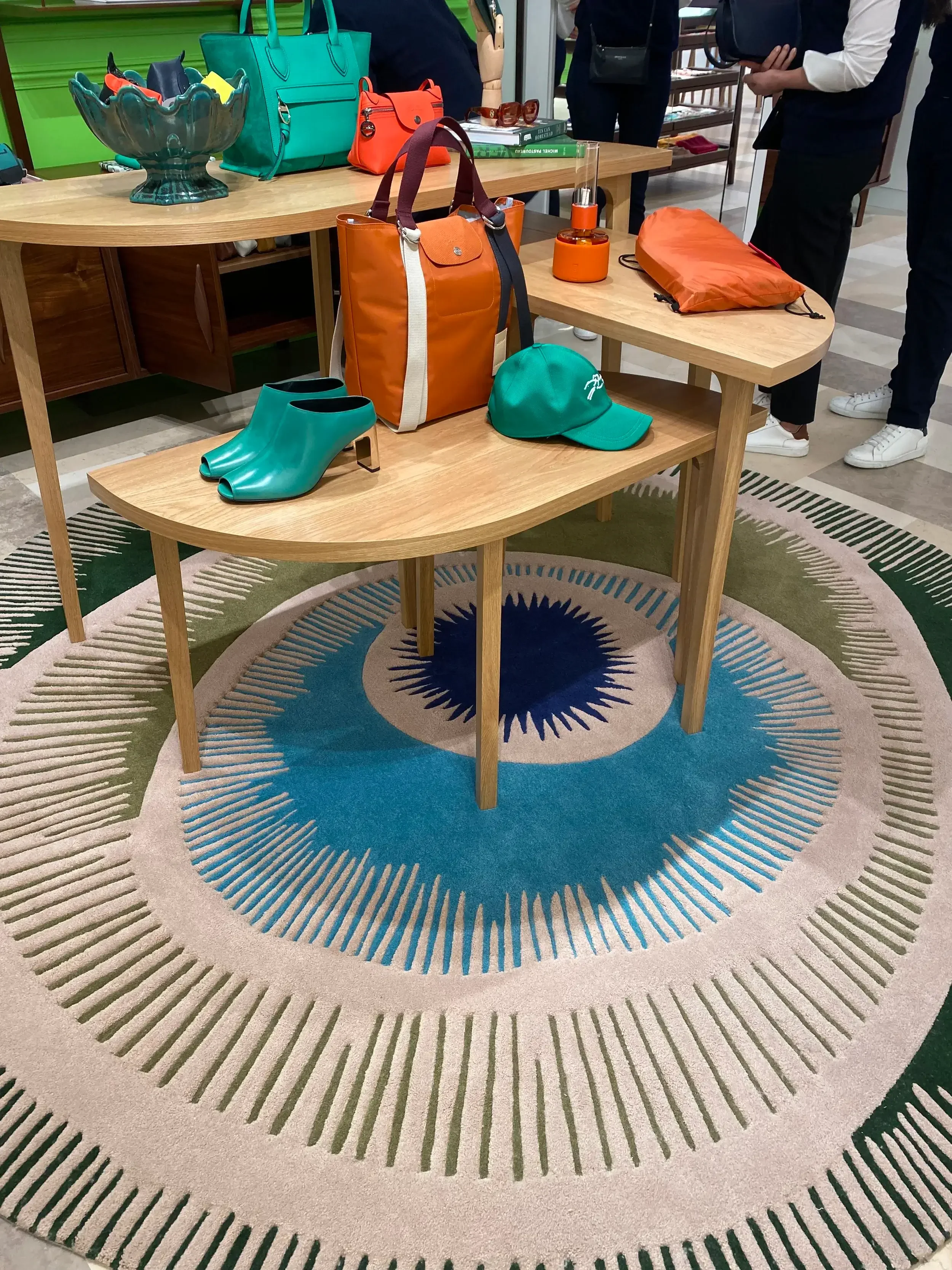 Table en bois avec plusieurs sacs à main colorés, une casquette verte, une paire de bottines vertes et une lampe orange, dans un magasin de mode