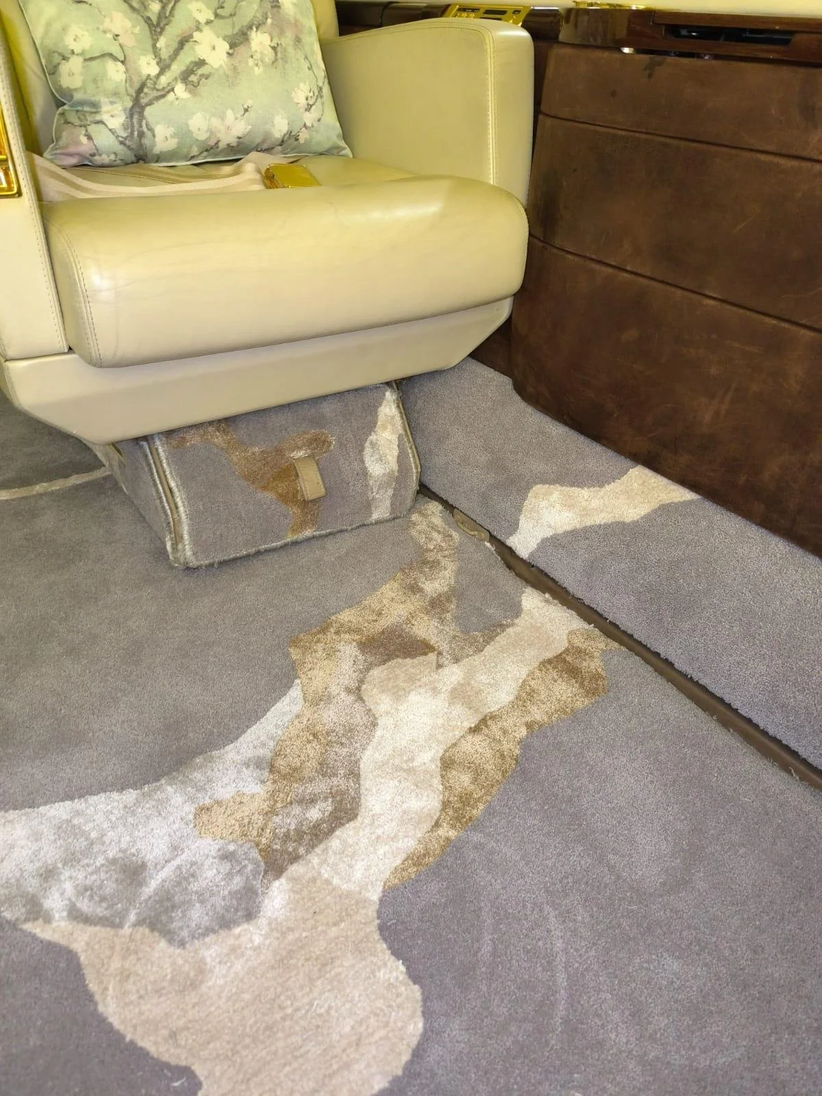 Tapisserie de sol déchirée, sous un fauteuil en cuir beige, avec une étagère en bois foncé à droite.