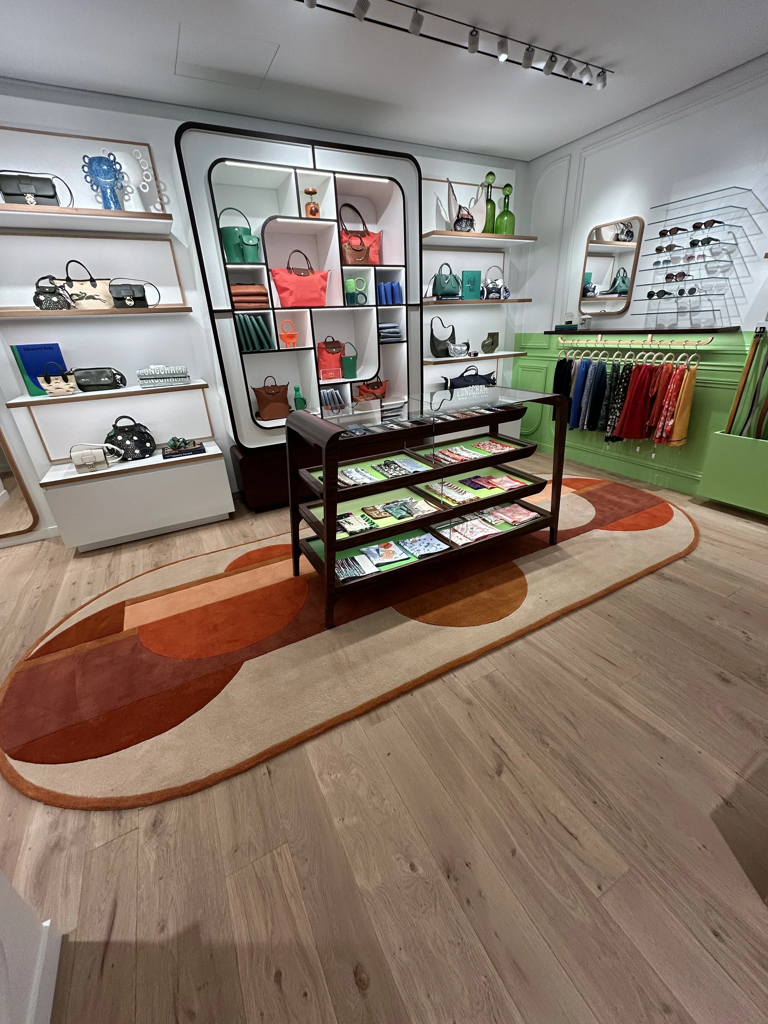 Une boutique de mode avec des sacs à main colorés, des vêtements suspendus et des accessoires exposés dans des étagères et sur un mur. Un tapis à motifs orange et beige couvre le sol en parquet.